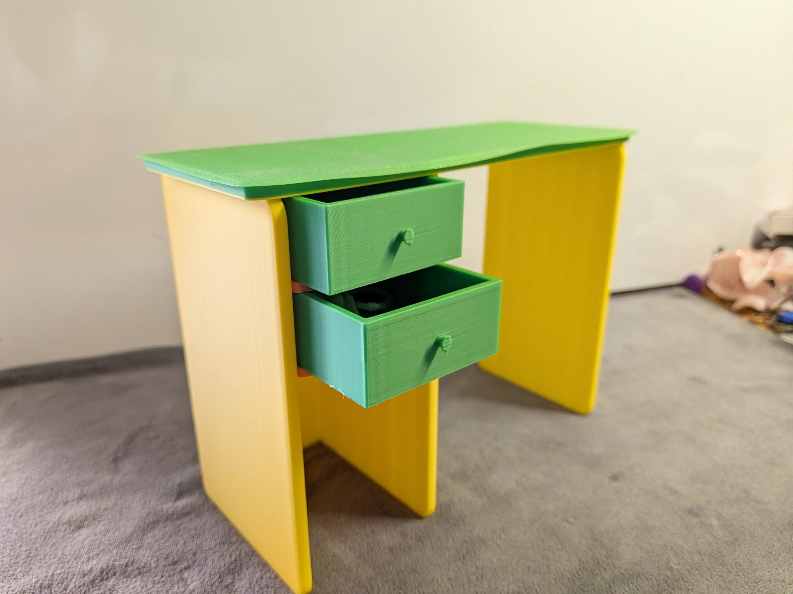 Modern 3D printed miniature dollhouse desk with a green top, yellow legs, and two functional green drawers pulled slightly open.