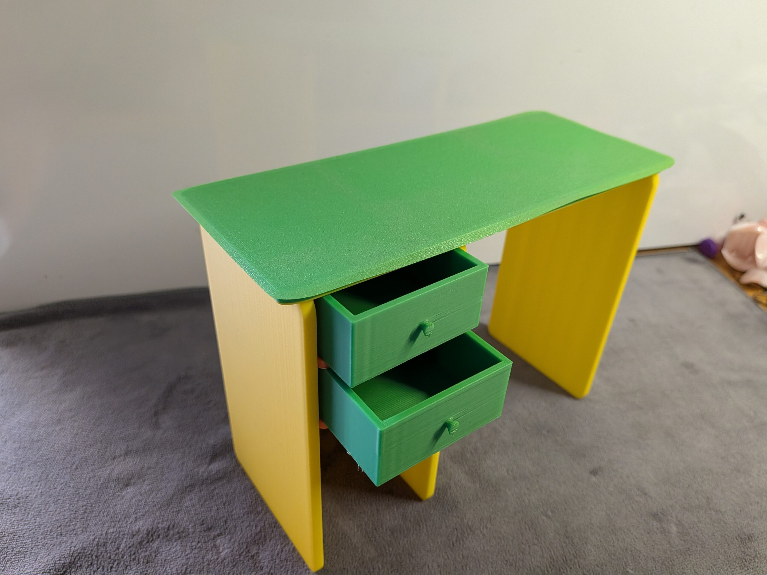 Modern 3D printed miniature dollhouse desk with a green top, yellow legs, and two functional green drawers pulled slightly open.