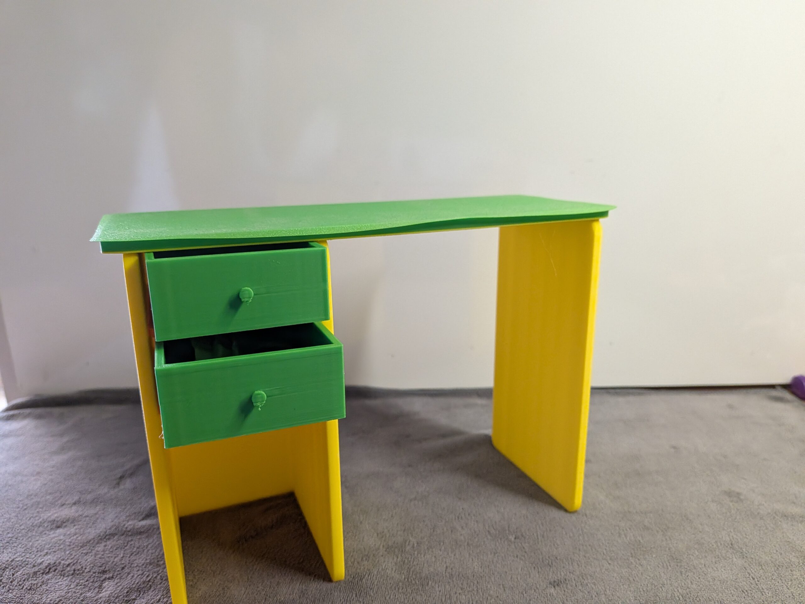 Modern 3D printed miniature dollhouse desk with a green top, yellow legs, and two functional green drawers pulled slightly open.