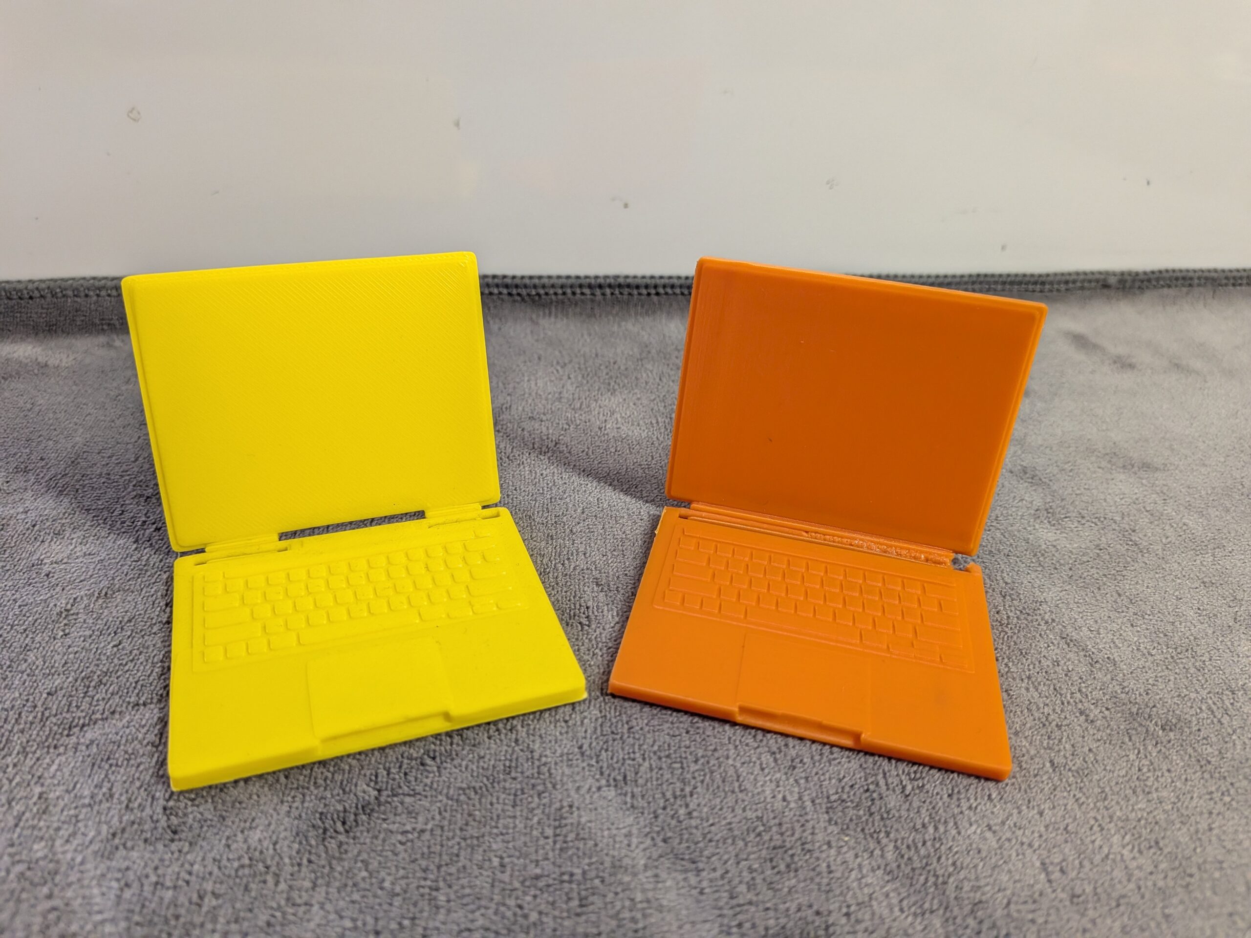 Modern 3D printed miniature laptops for dollhouses in yellow and orange with functional folding screens and detailed keyboards.