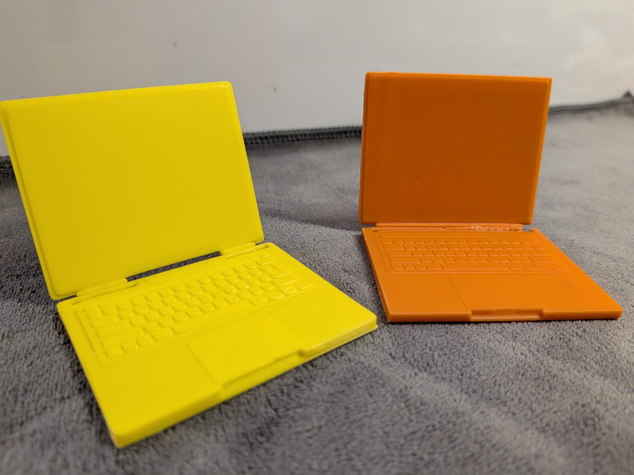 Modern 3D printed miniature laptops for dollhouses in yellow and orange with functional folding screens and detailed keyboards.