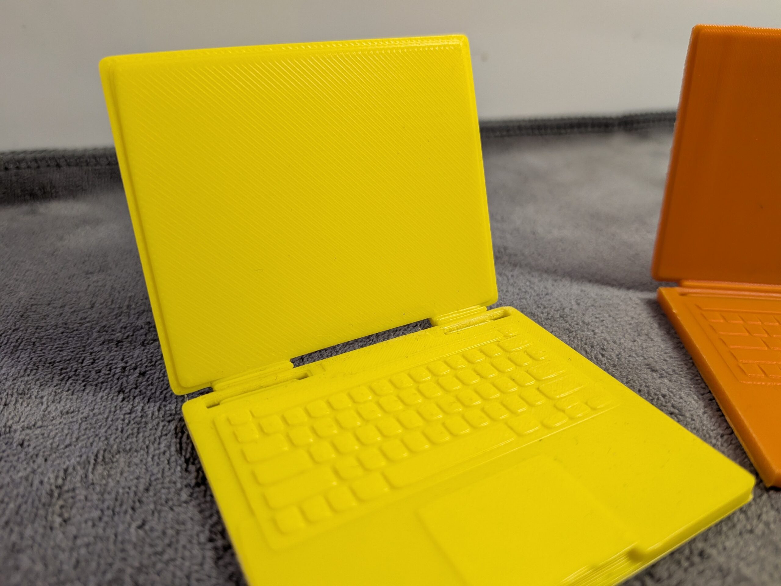 Modern 3D printed miniature laptops for dollhouses in yellow and orange with functional folding screens and detailed keyboards.