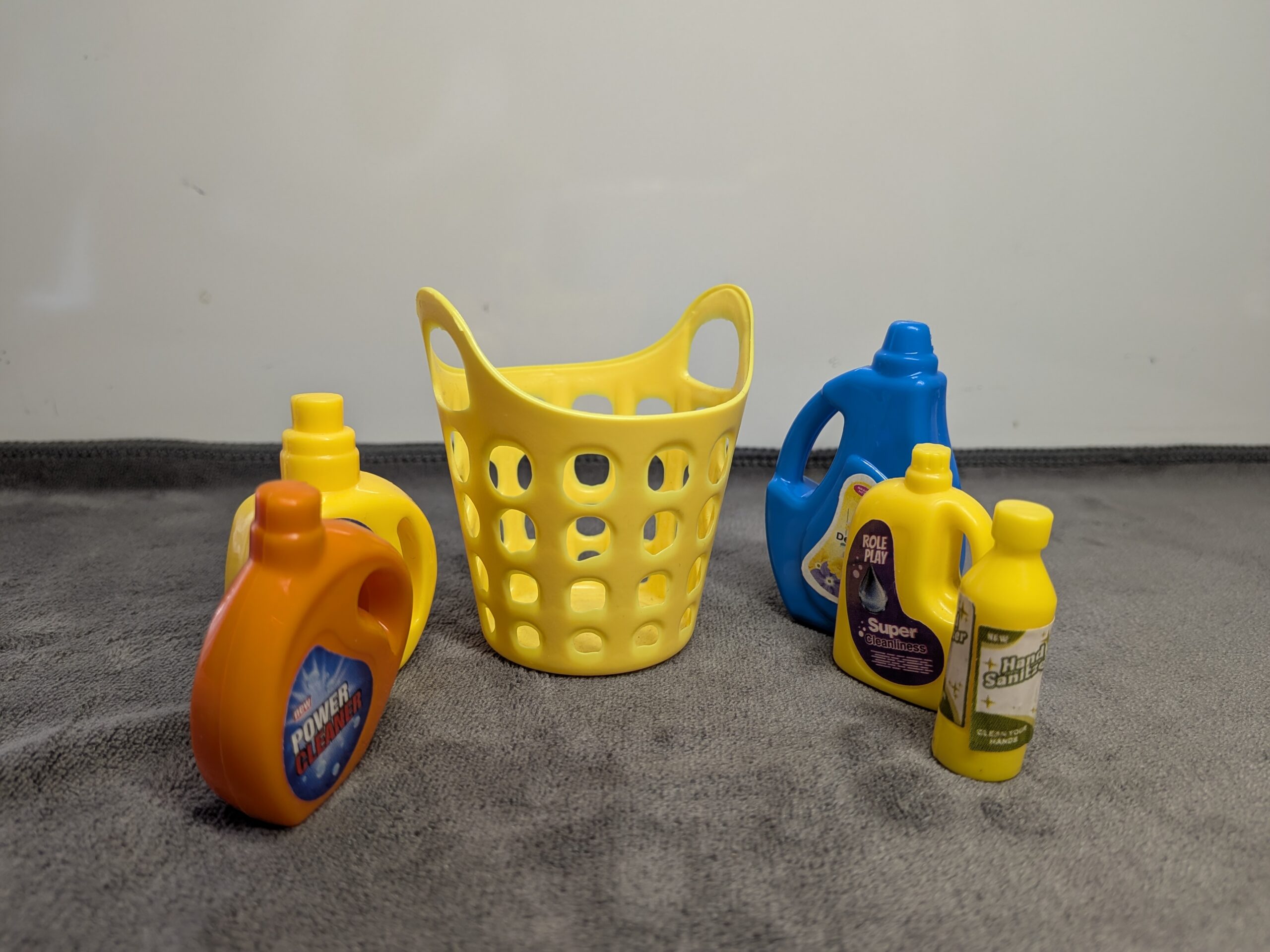 Assorted 3D printed miniature cleaning supplies including yellow, blue, and orange detergent bottles and a yellow laundry basket for a dollhouse.