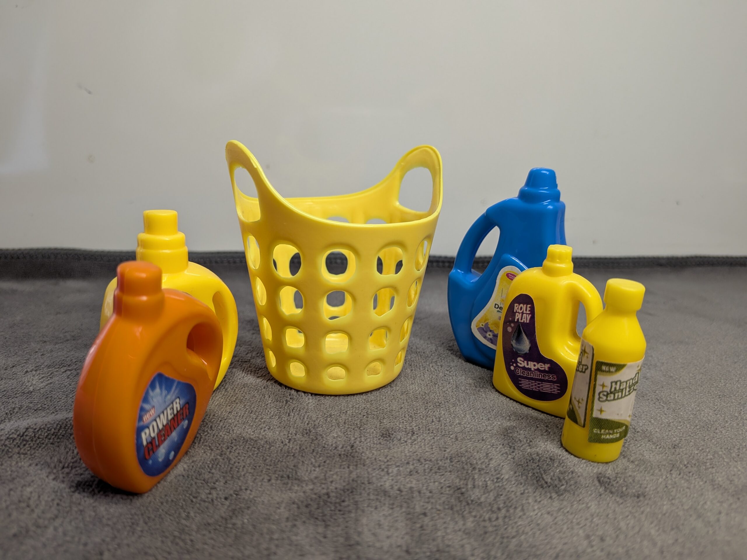Assorted 3D printed miniature cleaning supplies including yellow, blue, and orange detergent bottles and a yellow laundry basket for a dollhouse.