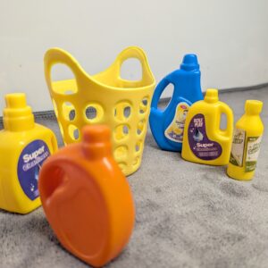 Assorted 3D printed miniature cleaning supplies including yellow, blue, and orange detergent bottles and a yellow laundry basket for a dollhouse.