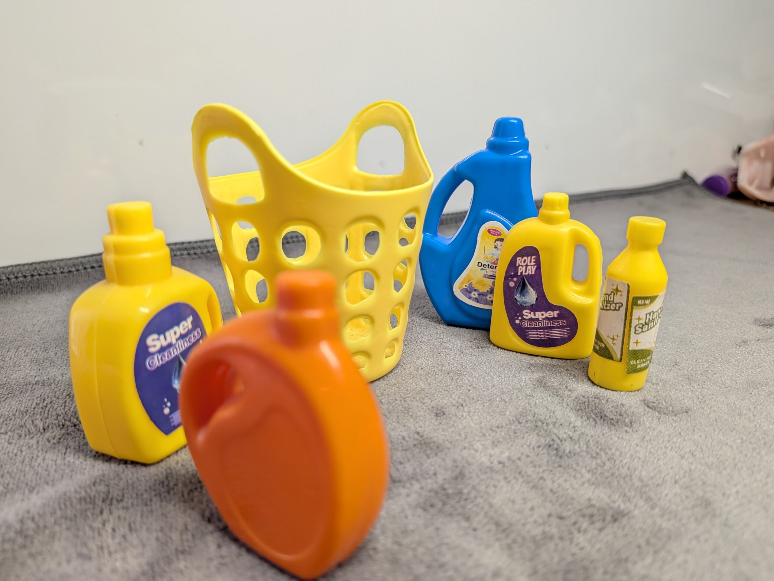 Assorted 3D printed miniature cleaning supplies including yellow, blue, and orange detergent bottles and a yellow laundry basket for a dollhouse.