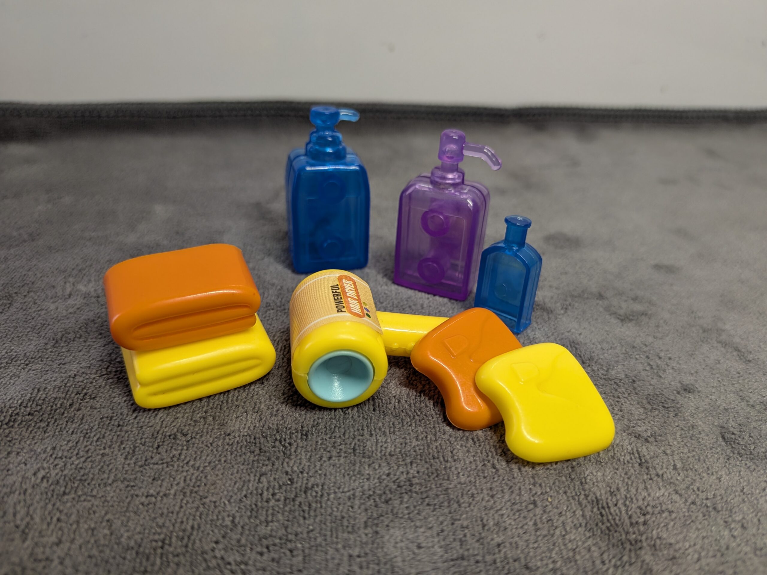 3D printed miniature bathroom set including translucent blue and purple shampoo bottles, a yellow hair dryer, and stacked orange and yellow dollhouse towels.