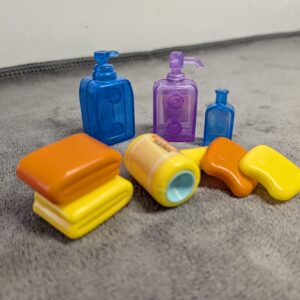 3D printed miniature bathroom set including translucent blue and purple shampoo bottles, a yellow hair dryer, and stacked orange and yellow dollhouse towels.