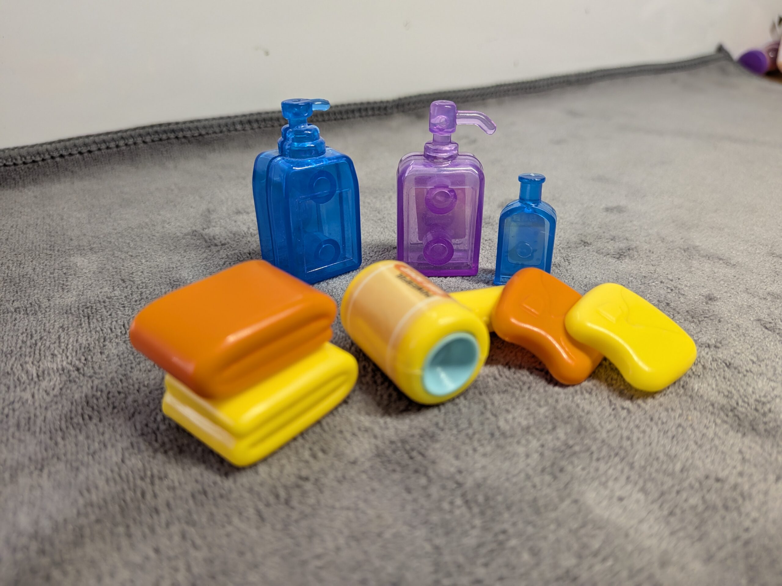 3D printed miniature bathroom set including translucent blue and purple shampoo bottles, a yellow hair dryer, and stacked orange and yellow dollhouse towels.