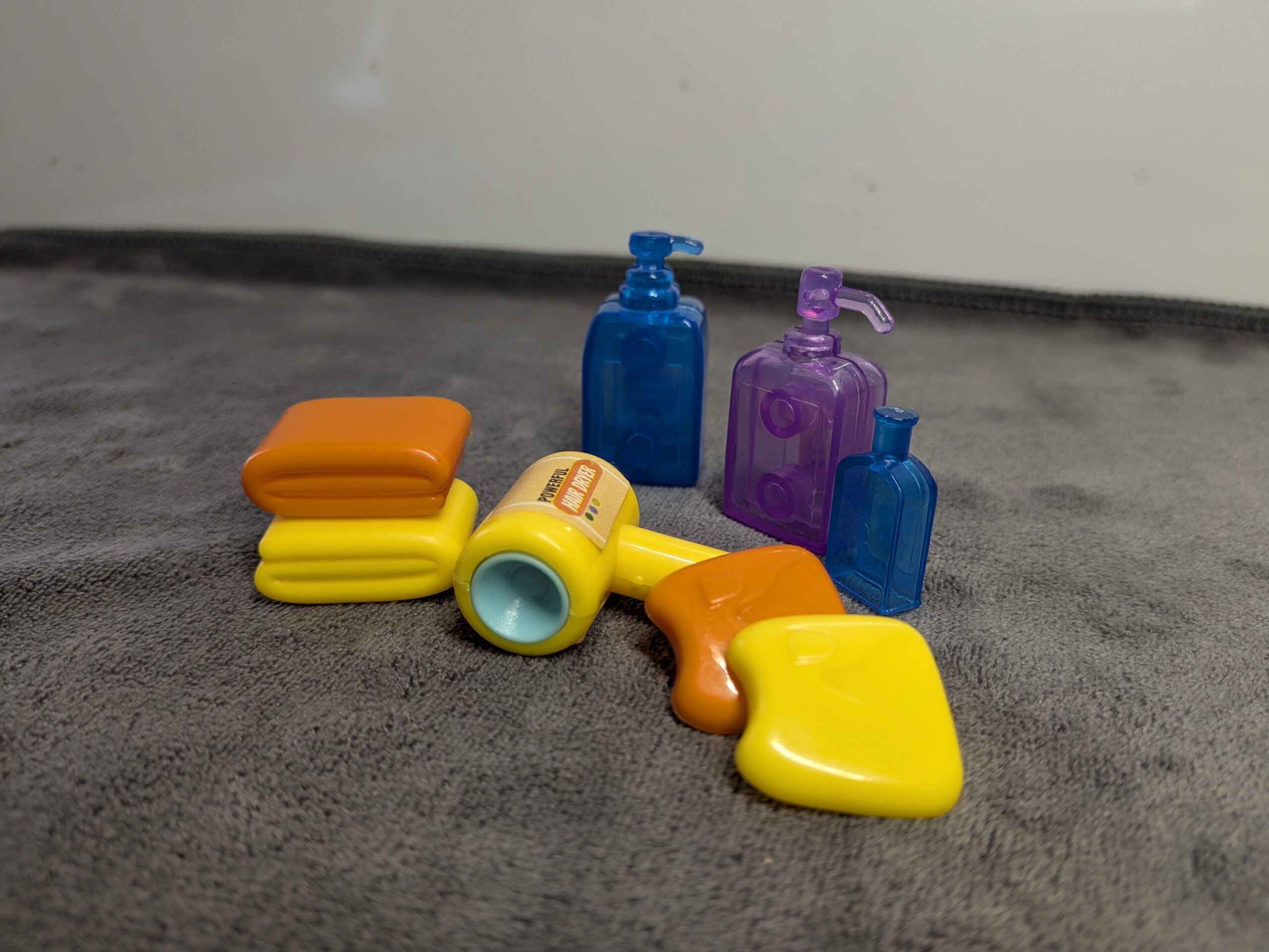 3D printed miniature bathroom set including translucent blue and purple shampoo bottles, a yellow hair dryer, and stacked orange and yellow dollhouse towels.
